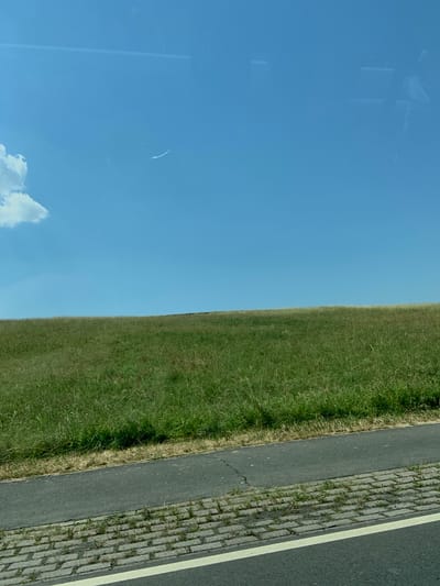 The image shows a grassy field next to a paved path with a cobblestone border and a white line. The sky is blue with some scattered clouds. The image is oriented vertically, with the sky occupying the left half and the field and path occupying the right half. There are reflections on the left side of the image, suggesting the photo was taken through a window. No people or specific objects are clearly identifiable. The time of day appears to be daytime, and the weather seems to be clear. The location context suggests this scene is in Niederwetz, Germany.