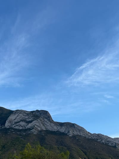 The image shows a mountain range in Ciudad Santa Catarina, Mexico (latitude: 25.658, longitude: -100.441). The mountain range is covered in vegetation. There are white streaks in the blue sky, indicating the presence of cirrus clouds. The image appears to have been taken on a clear day. The perspective is from a low angle, looking upwards at the mountain range. The image is well-lit and shows clear detail of the mountain range and sky.
