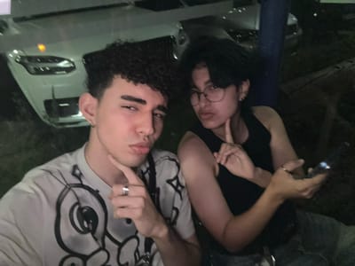 Two young adults pose closely together, one with dark hair and glasses wearing a black tank top and holding a phone, the other with curly hair and a white t-shirt with a graphic design. They are outside at night, and a silver vehicle is visible in the background. The image is rotated, causing the figures to appear sideways. Both individuals are looking at the camera with playful expressions.