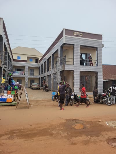 The image depicts an outdoor scene in Bokkos, Nigeria, likely during the daytime. A multi-story building with a modern facade, featuring horizontal gray and white patterned panels, dominates the left side of the frame. A sign on the building reads "RUKIN ASIRI PLAZA".
Several people are present, mostly young men. Some are standing near motorcycles parked on a dirt road in front of the building. One man in a red shirt is leaning towards a motorcycle, appearing to be engaged in conversation or interaction. Another man in a dark shirt and yellow shorts is also near a motorcycle, looking towards the viewer. A person in a red jacket is visible through a window on the second floor.
In the foreground, a stall is set up with various plastic containers and other goods for sale, including colorful tubs and jerrycans. The ground is a dusty, reddish-brown dirt, with some puddles indicating recent rain or dampness. The overall atmosphere seems to be that of a busy street or market area. The lighting suggests it's daytime, with an overcast sky. The image is oriented vertically, with the sky occupying a significant portion of the upper left.