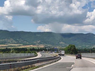 This image captures a highway scene near Dzhurovo, Bulgaria. The viewpoint is from a vehicle traveling on the highway. The road is multi-lane, with a concrete barrier separating the lanes and a metal guardrail on the left side. Several vehicles are visible, including a white truck with a trailer, a black SUV in the right lane, and other cars in the distance. The highway curves to the left, leading towards a landscape of rolling hills covered in dense green vegetation and patches of lighter colored fields. In the background, a large mountain range dominates the horizon under a partly cloudy sky with large white and gray clouds. A blue highway sign with white text, possibly indicating a destination, is visible on the right side of the road. The overall environment suggests a rural or semi-rural setting during daylight hours.
