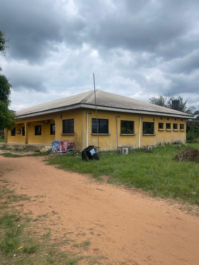 The image shows a two-story yellow building in Abraka, Nigeria, under a cloudy sky. The building has a corrugated metal roof, barred windows, and a tall metal pole extending from the roof. A black water tank is visible on the roof. Beside the building, there is a dirt road with green grass on the edge. A black trash can is positioned near the building, alongside colorful posters or fabrics. Trees and other vegetation are present in the lower left corner of the image. The scene appears to be outdoors in a rural or semi-rural setting.
