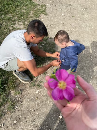 This high-angle photograph captures an outdoor scene featuring an adult, a young child, and a hand holding a flower. In the foreground, positioned to the right, a hand is seen holding a vibrant pink flower with a prominent yellow center, which is out of focus due to its closeness to the camera.

In the mid-ground, a man with short dark hair is seated or kneeling on the dirt ground, wearing a light grey t-shirt, light grey shorts, and olive green sneakers. He is facing a small child who is standing directly in front of him. The child, likely a toddler, is dressed in a dark blue hooded top with dark blue trousers. A small square patch on the child's top clearly displays the word "SKATE". The man's hands are gently interacting with the child's hands, suggesting an act of assistance or shared focus on an object between them. The child is looking downwards at their hands.

The ground beneath them is a mix of light brown dirt and gravel, with patches of green grass visible, particularly in the lower-left corner. Bright sunlight is evident, casting distinct shadows on the ground, indicating a clear, sunny day, likely midday or early afternoon. The scene conveys a gentle, caring interaction in an informal outdoor setting, identified as Пятый километр, Russia.