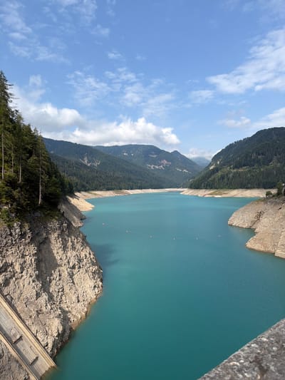 This is a vertical, eye-level, full shot of a reservoir in the Italian Alps, likely in the vicinity of Lateis / Latais. The turquoise water of the reservoir fills the center of the frame, stretching into the distance between tree-covered mountains. The banks of the reservoir are steep and rocky, with visible erosion and some vegetation. In the foreground, a concrete dam structure is partially visible on the left side of the image. A few small orange buoys are scattered across the water's surface. The sky above is blue with scattered white clouds, indicating fair weather. The scene is natural and serene, with no people or animals visible. The overall impression is one of a remote, mountainous landscape dominated by a large body of water.