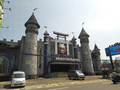 The image displays the exterior of a building designed as a themed castle, identified by signage as "NICOLE'S RIVER PARK," situated in the city of Ban Xieng Lom, Laos. The structure features a grey, stone-like facade with multiple turrets, each topped with a dark conical roof and an orange pennant flag. Blue structural elements accent the building, which includes small, yellow-framed windows. A large, reddish-brown teddy bear head is prominently mounted above the central entrance.
In the foreground, a paved road runs horizontally, separating the castle building from a parking area. A silver minivan is parked on the left side of the lot, facing the building. A dark grey minivan is parked further right, closer to the main entrance. To the right, a tall digital billboard displays colorful, illuminated graphics. Adjacent to the billboard, a large standing statue of a brown teddy bear, dressed in overalls, is visible. On the far right, a person wearing a helmet is riding a motorcycle on the road, partially obscured by the billboard's support structure. The scene is illuminated by daylight under a clear, pale blue sky, with visible overhead utility lines traversing the upper-left corner of the frame.