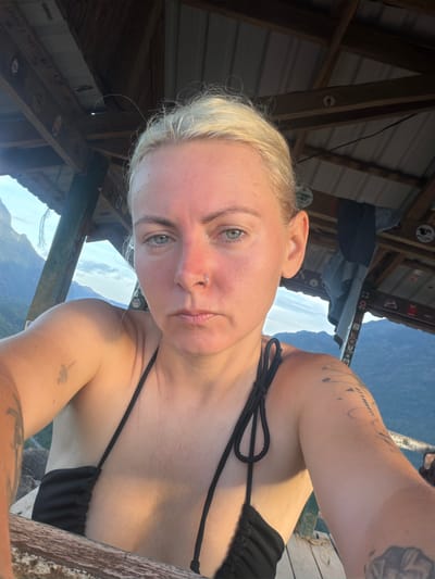 A woman with blonde hair and blue eyes, wearing a black bikini top, poses for a selfie in an open-air structure. She has a nose ring and several tattoos on her arms. The background shows a mountainous landscape with a cloudy sky, typical of the region around Naka, Laos. The structure appears to be made of wood and corrugated metal, possibly a lookout point or shelter.
