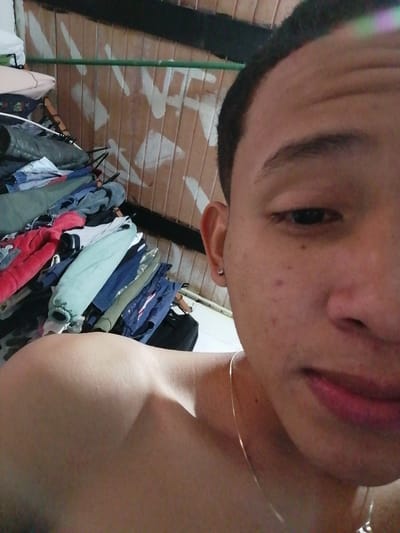 The image displays a close-up, chest-up view of a young individual, likely male, indoors in the city of Soacha, Colombia. The subject, positioned slightly off-center to the right, has short dark hair with a faded hairline, dark eyes, and a small silver stud earring in the right earlobe. Visible facial features include a prominent right eyebrow, minimal facial hair, and several small blemishes on the right cheek. The subject's right shoulder and clavicle are visible, suggesting they are shirtless or wearing light attire, and a thin silver-colored chain necklace hangs around the neck. The subject is looking directly at the camera.
The background on the left side features a substantial, disorganized pile of various clothing items, including jackets and shirts, in colors such as red, light blue, dark blue, grey, and white, stacked on an unseen surface. Behind the clothing, parts of a dark-colored bag are visible. The background on the right, behind the subject's head and shoulder, consists of a light brown or beige wall, possibly wood paneling, featuring abstract white spray-painted markings. A thin green horizontal bar runs across the wall above these markings, and a dark horizontal trim piece is visible at the top edge of the frame. The lighting appears artificial, illuminating the subject from the left.