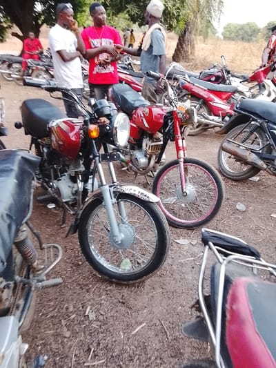 An outdoor scene in Tabu, Nigeria, depicts a gathering of individuals amidst numerous motorcycles on a reddish-brown dirt ground. Three adult males are engaged in a central interaction. The man on the left wears a white t-shirt and sunglasses, holding his left hand near his face. The central man wears a red t-shirt with "DEY RIDE" text and gestures with both hands. The man on the right, older, wears a light blue shirt, a light cap, and has a light fabric draped over his shoulder; he faces the central man. Behind them, other individuals are visible, including one in red seated on a motorcycle on the far left.
A cluster of motorcycles, predominantly red, occupies the mid-ground and background. In the immediate foreground, two red motorcycles are prominent. The one on the left has a patterned red tank, black seat, chrome headlight, orange indicator, and a silver front fender with partial text "JUST TRUL". Its front wheel has a black tire and silver spokes. The motorcycle to its right also has a red frame and tank, a chrome headlight, and a distinctive pink rim on its front wheel, with a black tire. Other red motorcycles are visible further back, including one with "JAGGAD" text on its side panel and another showing a dark, corroded exhaust pipe. The environment includes a large tree with green foliage behind the central figures and dry vegetation in the distance, under a bright sky.