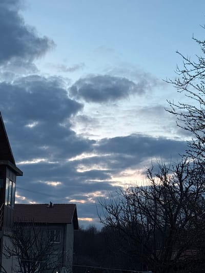 The image captures an outdoor scene in Zidartsi, Bulgaria, at an unspecified time between dusk and dawn. The sky, occupying approximately the upper two-thirds of the frame, is predominantly covered by a layer of dark gray, cumuliform clouds. Horizontal streaks of a lighter, illuminated sky, exhibiting pale yellow and blue hues, are visible through breaks in the cloud cover, particularly towards the horizon.
In the lower-left quadrant, a partial view of a multi-story residential building is present. Its dark-eaved roof slopes downwards from the upper-left. Below the roofline, a section with multiple reflective windows, possibly an enclosed balcony or sunroom, reflects the sky. A dark metallic railing or support structure is visible below the windows.
To the right and slightly behind this building, a second, two-story house is visible. It features a prominent reddish-brown tiled gable roof. A solitary, dark-colored avian figure is perched centrally on the ridge of this roof. The house's exterior walls are a muted gray, and a single rectangular window is visible on the upper floor.
Bare, deciduous tree branches, devoid of leaves, are silhouetted against the sky, framing the right side of the image from top to bottom and extending across the lower mid-section. A dense, dark line of trees or forest forms the background horizontally, positioned behind the structures and foreground foliage. Faint, dark utility lines are discernible stretched horizontally across the mid-ground, above the second house's roof. No human subjects or active interactions are observed.