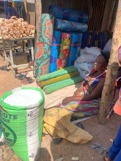 The image depicts an outdoor market stall in Nigeria. The primary subjects are a woman, likely a vendor, and various goods for sale. The woman is wearing a brightly colored, patterned dress and appears to be resting or attending to her merchandise.
The objects for sale include an assortment of rolled-up mats, some with colorful patterns and cartoon characters, others with striped designs. There are also several large sacks, one of which is predominantly green and white and contains a white granular substance, possibly fertilizer given the visible text on it. Other sacks are a pale yellow color and appear to be made of woven material. A pile of small, brown, individually packaged food items, possibly snacks or sweets, is also visible, neatly arranged on a shelf. A pair of sandals lies on the ground near the woman. The setting is rustic, with corrugated metal sheeting visible in the background and loose gravel or dirt on the ground. A wooden beam runs overhead, forming part of the stall's structure.
The time of day appears to be daytime, with natural light illuminating the scene. The weather seems to be warm, typical of an outdoor market in Nigeria. The overall impression is one of an active, albeit somewhat cluttered, marketplace.
Visible text on the green and white sack reads: "DANGOTE FERTILIZER" and "ER" at the bottom edge. The phrase "DANGOTE FERTILISER" is repeated in smaller print along the top band of the sack.