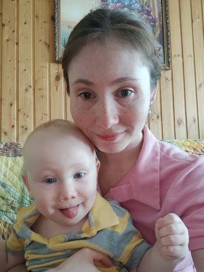 A woman and a baby are posed closely together for a selfie. The woman, with fair skin and freckles, wears a pink collared shirt and earrings. Her hair is pulled back. The baby, also fair-skinned with blue eyes, sticks out its tongue. The baby wears a yellow and grey striped collared shirt. They are sitting on a patterned couch or chair. Behind them is a wooden wall with a framed painting of a landscape hanging on it. The location is suggested to be Malaya Purga, Russia.