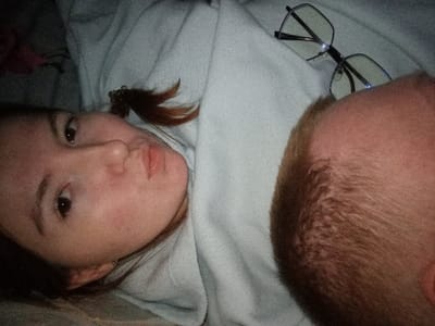 This image depicts two individuals resting on light gray fabric, presumably bedding, in an indoor setting within the city of Старый Шагирт, Russia. The female subject, positioned on the left side of the frame, has fair skin, dark eyes, and dark hair secured with a brown hair tie, creating a small ponytail. She is wearing a light gray hooded garment and looks directly towards the viewer with a neutral expression. Adjacent to her on the right, the top of a male's head is visible, showing short-cropped, light brown hair. A pair of black-framed, rectangular eyeglasses with lenses reflecting a blue light sheen rests on the light gray fabric, positioned between the two subjects and near the male's head. A dark, indistinct object is partially visible in the upper left background.