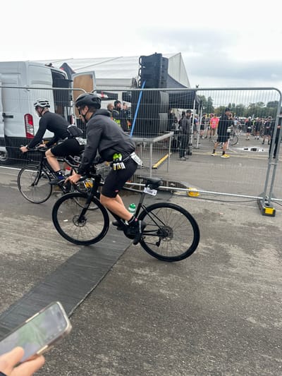 The image depicts an outdoor cycling event taking place on a paved surface under an overcast sky in Weymouth, United Kingdom. In the immediate foreground, a person's left hand holds a smartphone, partially obscuring a dark, wide strip on the asphalt ground, possibly a timing mat.
Two male cyclists on black road bikes are central to the midground, moving from left to right. The lead cyclist wears a black helmet, sunglasses, a dark grey long-sleeved top or jacket, black cycling shorts, and black cycling shoes with "Specialized" branded socks. A green water bottle is in a cage on the bike frame, and a race number "1888" is attached to the seat post. A yellow-green pouch and white papers are affixed to the rider's lower back. The second cyclist, positioned slightly behind and to the left, wears a white helmet, a black long-sleeved top, black cycling shorts, and black cycling shoes, with a race bib visible on their lower back. Both cyclists are actively pedaling.
To the left, behind the cyclists, the rear of a white utility van with its doors open is visible, adjacent to a large white marquee tent. To the right of the cyclists, a metal barrier system separates the cycling track from an event area. Behind these barriers, a stage structure is equipped with multiple stacked black speaker units. Various individuals are present in this background area; one person in a yellow high-visibility vest stands near the speakers, and another, further right, appears to be holding a microphone. A crowd of participants, many in cycling attire and helmets, are gathered with bicycles in what appears to be a staging or waiting area. Small puddles are visible on the paved ground in this area. The far background shows trees beneath the overcast sky.