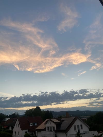 The image depicts a dusky sky over a residential area, likely in Kassel, Germany. The sky is filled with wispy, orange-tinted clouds in the upper portion, suggesting either a sunset or sunrise. Below these, a band of darker, more textured clouds stretches across the horizon. In the foreground, the rooftops of several houses are visible. One house in the center has a prominent red-tiled roof with solar panels and dormer windows. Adjacent houses also feature tiled roofs and residential architectural elements. The scene is framed by trees and a distant, rolling landscape with faint lights indicating buildings. There are no people or animals visible.