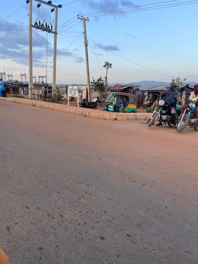 The image captures a vibrant street scene in Nabatum, Nigeria, likely during the late afternoon or early evening under a light blue sky with scattered clouds. The primary focus is a paved road that curves slightly to the left, flanked by an unpaved shoulder and various structures.
On the right side of the road, several individuals are gathered near and on motorcycles. One man, wearing a blue shirt and a dark cap, is seated on a motorcycle parked on the shoulder. Another motorcycle is visible directly behind him. Further along, numerous informal shops or stalls with corrugated metal roofs line the roadside, indicating a commercial area.
On the left side of the road, a yellow and green trishaw (motorized tricycle taxi) with the license plate "PTA-0285" is prominently parked. Overhead, multiple utility poles with complex wiring stretch across the frame, carrying power lines. In the background, beyond the immediate structures, distant mountains or hills are visible under the soft light.
A white sign, partially obscured and rotated, is visible on the right side of the road near the utility poles. Text on the sign includes "SOLANTO GROUP OF COMPANIES CORPORATION" and further details such as "P.O BOX", and potentially locations like "KADUNA" and "ABUJA", suggesting business operations. The asphalt road surface shows visible texture and speckles. The scene depicts typical daily life and commerce in a Nigerian urban or semi-urban setting, characterized by local transportation, informal businesses, and utility infrastructure.