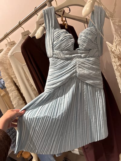 A blue pleated dress hangs on a clothing rack among other garments. The rack is located in an unidentified retail environment in the city of Sunrise, United States of America. The garments on the rack include a brown top, a white sweater, and a lacy white garment. A hand is seen holding up the edge of the blue dress. The hangers are marked with "cult gaia".