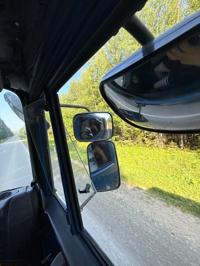 This is a first-person perspective, POV, shot from inside a vehicle, possibly a tractor, looking out through the driver's side window and mirrors. The scene depicts a rural road with lush green vegetation and trees on either side. The road is paved and appears to be a two-lane highway. The interior of the vehicle is dark blue and some of the dashboard and controls are visible. The mirrors reflect the surrounding environment and the interior of the vehicle. In the side mirrors, the reflections show a person holding a phone, possibly taking the photo. The time of day appears to be daytime, with clear skies and sunlight filtering through the trees. The location is described as Откормочная ферма, Russia, which is a rural area consistent with the imagery.