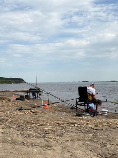 The image shows a man fishing on the edge of a body of water. He is seated in a specialized fishing chair with equipment, including multiple fishing rods set up along the bank. The ground consists of dirt and dry reeds or grass. The water is calm, and the sky is partly cloudy. The scene appears to be in Krasnyi Bor, Russia, based on the information given.