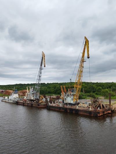 The image captures a scene at a port in Vytegra, Russia, on an overcast day. The foreground is dominated by dark, rippling water. In the mid-ground, a series of connected barges are moored. Two large cranes are prominent on these barges; one is a taller, yellow crane with a visible operator's cabin and a hook at the end of its boom, and the other is a gray crane with a similar structure, also equipped with a hook. Several other pieces of heavy machinery, possibly also cranes or excavators, are visible on the barges, some appearing weathered and rusty.
A white vessel, possibly a tugboat, is docked at the front of the barges, and a large pile of logs is stacked on the shore behind it. The background is a dense line of green trees, indicating a forested area. The sky is a muted gray with visible cloud formations. The overall impression is one of industrial activity in a natural setting. There are no visible people in the image.