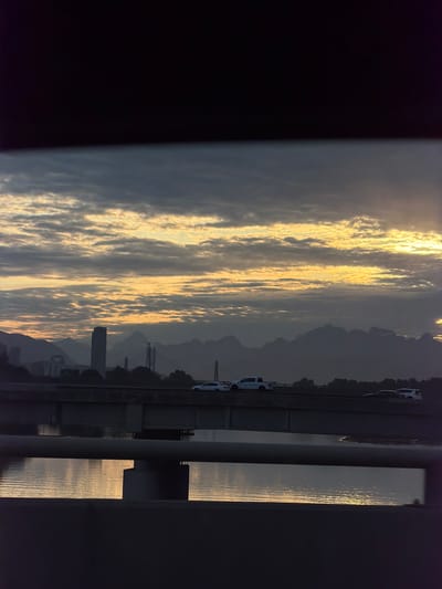 The image shows a dusky scene in Delta, Canada, likely at sunset. The view is framed by dark structures, possibly a bridge or building, on the left and right sides. In the center, a body of water reflects the golden light of the setting sun, creating shimmering patterns on the surface. Beyond the water, silhouetted mountains rise in the distance, adding depth to the landscape. A line of cars can be seen, indicating a road or bridge. In the middle distance, building-like structures stand out in the cityscape. Overall, the scene conveys a sense of tranquility and beauty in the Canadian landscape.