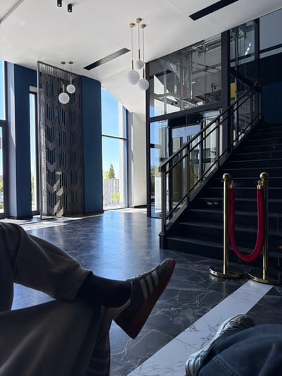 The image shows an interior view, apparently within a modern building located in Verin Ptghni, Armenia. The space features a contemporary design with a high ceiling, accented by modern globe lighting fixtures. A decorative geometric panel is on the wall. There is a glass-enclosed elevator shaft. A staircase is present, demarcated by gold stanchions and red velvet ropes. The flooring has a marble-like pattern in dark and light tones. A pair of legs, one crossed over the other, is visible, with a sneaker and a brown-soled shoe on the feet. A person is sitting in the foreground. A partial view through windows reveals a glimpse of greenery.