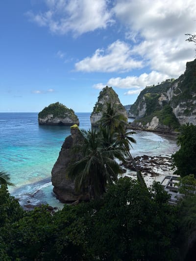 The image showcases a tropical coastal scene, likely in Pejukutan, Indonesia, given the limestone cliffs and turquoise waters. Two prominent rock formations, one with palm trees, are visible in the foreground. The water transitions from a deep blue further out to a vibrant turquoise closer to the shore. A rocky beach is present in the distance. Dense, green vegetation frames the right side of the image, while the sky is filled with white, fluffy clouds against a blue backdrop. Part of a man-made structure is visible above the rocky beach.