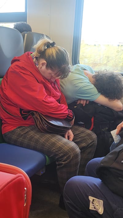 On what appears to be a train traveling through Lukhovitsy, Russia, a woman with blonde hair pulled back in a ponytail sleeps in a red jacket. She is seated next to a person with curly hair wearing a light blue shirt, who is also sleeping against a black backpack. Another person wearing blue pants with a patch is partially visible on the right, and a red suitcase is visible in the lower right corner. The train window shows an outdoor scene with trees. The people are seemingly sleeping.