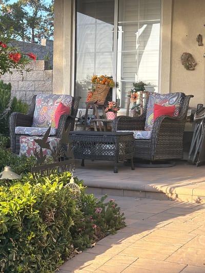 This image depicts an outdoor patio area at a residence in Aliso Viejo, California, USA. The scene is set on a paved patio with tan-colored stone tiles. In the background, a sliding glass door with white blinds is visible, reflecting sunlight. To the left of the door, a portion of a building wall made of light beige stucco is present, with decorative items attached, including a piece of driftwood with plants.
The foreground features two wicker rocking chairs with patterned cushions and red accent pillows. Between the chairs sits a dark wicker coffee table. In front of the chairs, a metal yard decoration with bird silhouettes and the word "WELCOME" is partially obscured by lush green foliage. A birdcage decoration hangs from the foliage on the right. Red and orange flowers are visible in the upper left corner of the image, cascading from a planter. A tiered wooden stand with potted plants and flower decorations is positioned between the chairs and the sliding glass door.
The lighting suggests it is late afternoon or early evening, with long shadows cast across the patio. The environment appears to be a residential backyard, with a stone wall and landscaping visible in the background.