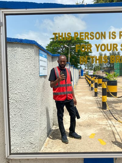 The image depicts a man standing in what appears to be an industrial area or a port. He is wearing a black t-shirt, black pants, and a high-visibility red vest over his t-shirt. He is holding a smartphone in his right hand and a dark-colored object, possibly a rolled-up umbrella or a bag, in his left hand.
The man is positioned in front of a reflective surface, likely a large pane of glass or a polished metal surface, which reflects the environment behind him. The reflection shows a pathway lined with yellow and black striped bollards and chains, suggesting a restricted or designated area. In the background, there are shipping containers stacked high and trees. The sky is visible with some clouds.
A sign with yellow lettering is visible on the reflective surface, reading "THIS PERSON IS R FOR YOUR S IN THIS BR". The lettering is partially obscured and appears to be in English.
The environment is outdoors, with a concrete ground. There are yellow markings on the concrete, possibly indicating spills or warnings. The wall on the left of the image is textured and painted white, with a blue painted border at the top. Another sign with text in what appears to be a reversed script (due to the reflection) is mounted on this wall.
The overall impression is a scene within a port or industrial facility, where safety regulations are likely in place, as indicated by the high-visibility vest and the bollards. The location is identified as Kinkinau, Nigeria.