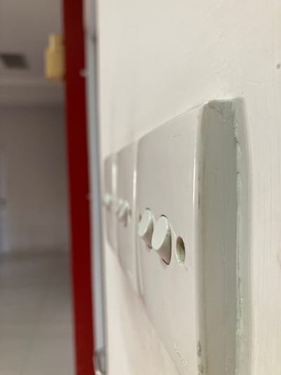 A close-up, eye-level shot focuses on a row of white, modular light switches mounted on a white wall. The switch on the right is most in focus, revealing two round, white buttons protruding from its face. The wall exhibits slight wear and tear, with some scuff marks and a faint crack near the corner of the switch plate. To the left of the switches, a red door frame is visible, partially obscuring an out-of-focus interior. The background is indistinct, showing muted colors and suggesting a hallway or room. The scene is devoid of people. The location is not specified in the image or its context.