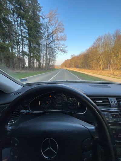 The image presents a static, first-person perspective from the driver's seat of a Mercedes-Benz vehicle, situated on a rural road within the city of Weißenhorn, Germany. The vehicle's interior components are prominent in the foreground, including a black steering wheel bearing the Mercedes-Benz logo, and an instrument cluster. The instrument cluster shows a speedometer needle resting at 0 km/h, a digital display indicating "0 km/h" and "0.0 l/100km," and an odometer reading of "6271.9 km." An amber warning light is illuminated on the dashboard. Further right, a portion of the infotainment system displays "24°."
The view through the windshield reveals a straight, two-lane asphalt road with a broken white centerline and a solid white line marking the right edge. Parallel to the right side of the road is a narrower paved path. The road is bordered by natural vegetation: a dense line of tall, slender coniferous trees forms a forest edge on the left, while a line of deciduous trees, mostly bare of leaves, flanks the right side of the road and path. These bare trees exhibit a warm, golden illumination consistent with direct sunlight. In the distant midground, two small, indistinct human figures are visible on the paved path, appearing to walk away from the viewer. The sky above is clear and bright blue, devoid of clouds.