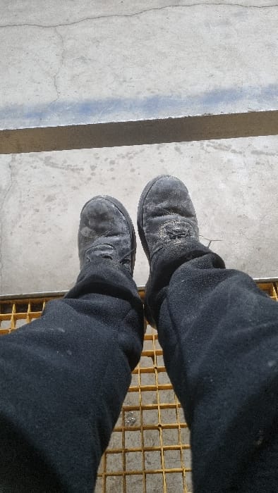 The image, taken from a first-person perspective looking down, shows a person's legs and feet. The individual is wearing dark, possibly black, trousers that appear somewhat dusty or worn, and black work boots. The boots are visibly scuffed and covered in a light layer of dirt or dust, indicating heavy use.
The ground beneath the person consists of a concrete surface, which also shows signs of dirt and some cracks. A light blue painted line runs across the concrete from left to right, creating a distinct boundary or marker. To the right side of the frame, underneath the person's right foot, a yellow metal grate is visible, suggesting a drain, platform, or industrial flooring. The lighting indicates it is daytime. There are no specific cues for weather, emotions, or visible text within the frame. The overall scene suggests an outdoor or industrial environment. The location context provided is Chapyk, Uzbekistan.