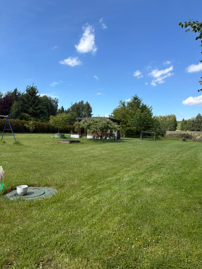 A wide-angle, eye-level, daytime, outdoor shot shows a lush green lawn in a backyard in Strzyżowice, Poland. The sky is bright blue with a few scattered white clouds. On the left side of the frame, a blue swing set stands partially visible. In the middle ground, a wooden gazebo with a thatched roof is nestled among trees. Several chairs and a table are visible under the gazebo. A small sandbox is located in front of the gazebo. A green soccer goal with a net is positioned on the right side of the lawn. Various trees and hedges surround the property, with a line of denser trees forming a backdrop at the rear of the yard. In the foreground, a round, green access hatch is visible on the grass, with a white ceramic pot placed on top of it. The grass is well-maintained and dotted with small white flowers.