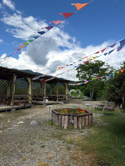 The image depicts an outdoor seating area with wooden gazebos and picnic tables, decorated with colorful bunting strung across the sky. In the foreground, a wooden planter box contains vibrant red and yellow flowers. Beyond the gazebos, a lush green hillside is visible, with a large tree and a heart-shaped floral arrangement in the distance. The sky is partly cloudy with scattered white clouds against a bright blue backdrop. The ground is covered with gravel and some patches of grass, with a stone path winding through the area. The setting appears to be a recreational or picnic spot in Izmaylovka, Russia. There are no people visible in the image.