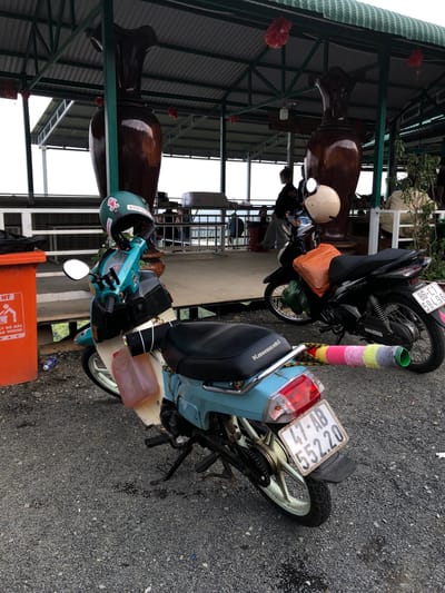 In Nam Hải, Vietnam, under a covered outdoor area supported by green metal beams, two parked motorbikes are visible on a gravel surface. The motorbike in the foreground is a light blue and cream colored scooter with a green helmet adorned with stickers placed on its handlebars. A translucent pink plastic container is attached to the side of this motorbike. A roll of colorful yarn, in shades of pink, yellow, and gray, is secured to the rear of the scooter. The motorbike in the background is black and has an orange plastic bag and a green plastic bag strapped to it. A beige helmet rests on its handlebars. Large, dark brown decorative vases are positioned behind the motorbikes. In the background, there are white tables and chairs and a railing overlooking an unseen area. An orange trash receptacle with Vietnamese text and a graphic of a person discarding waste is situated to the left of the foreground motorbike.