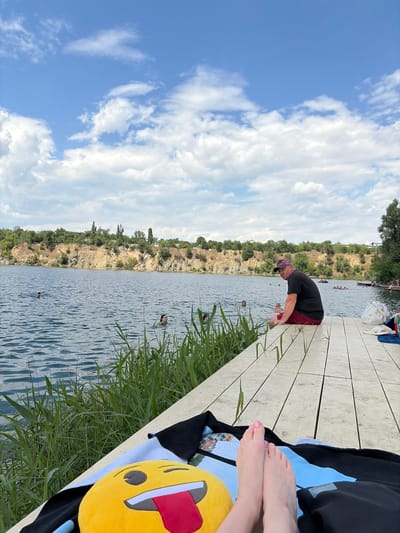 In this image, several people are enjoying a sunny day by a lake. A man wearing a red cap and a black t-shirt is leaning over a wooden dock, possibly about to enter the water. In the distance, other individuals are swimming in the lake, and some appear to be on small boats. The lake is surrounded by a rocky, tree-covered embankment. The sky is a clear blue with fluffy white clouds, suggesting fair weather.

In the foreground, on the right side of the dock, a person's feet are visible, resting on a blue towel. A bright yellow emoji pillow with a red tongue sticking out lies on the towel. The overall scene depicts a recreational activity at a natural lake, likely during daylight hours given the bright sky and shadows. The atmosphere seems relaxed and summery. The location is identified as Novomykolaivka, Ukraine.