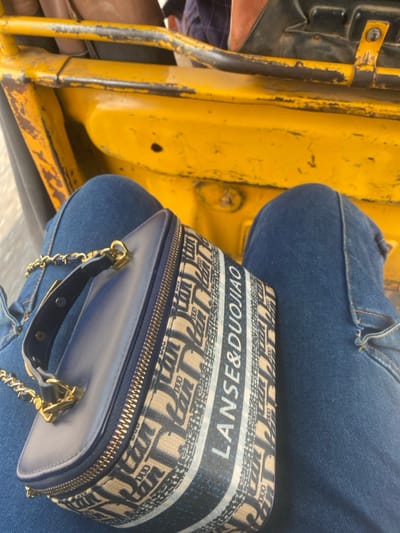 The scene depicts the interior of a worn yellow vehicle in Odo Ona, Nigeria. An individual is seated, with their lower legs, clad in blue denim jeans, prominently visible. A rectangular handbag rests on the individual's lap, positioned between the knees. The handbag is characterized by a beige and dark blue fabric pattern featuring stylized 'D' motifs and the text "LANSE & DUOJIAO". It has a dark blue top section, a gold-colored zipper closure, a small dark blue handle, and a gold-colored chain strap. The vehicle's interior structure consists of yellow-painted metal components, exhibiting visible wear, scratches, and rust.