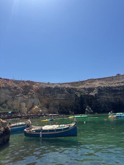 The image depicts a sunny day at a coastal area in Mellieħa, Malta. Two traditional Maltese boats are moored in the clear, turquoise water in the foreground. The boat on the right is blue and white with a cream-colored cover on its stern, while the boat on the left is primarily blue with white trim. Behind the boats, a rocky cliff face rises, exhibiting layers of sediment in shades of brown and tan, with some sparse green vegetation. A metal fence runs along the top of the cliff. In the middle ground, several inflatable water toys, including a yellow lily pad and green platforms, are scattered in the water, with people visible interacting with them and swimming. Further back, a wooden pier or boardwalk extends into the water, with small wooden structures that appear to be shelters or changing rooms. The sky is a clear, bright blue, indicating a cloudless day.