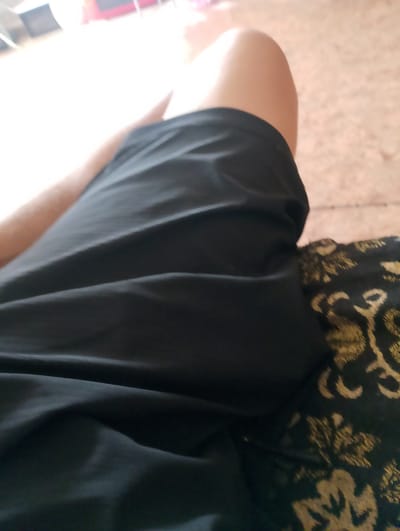A close-up, top-down shot shows a person's legs and thighs covered by black fabric. The fabric is draped over the left leg and thigh, with the right thigh exposed and slightly curved. The background is a blurry, beige surface with a patterned texture. The overall lighting is dim. The scene is indoors, possibly a home or a casual setting in Лобаново, Kazakhstan.