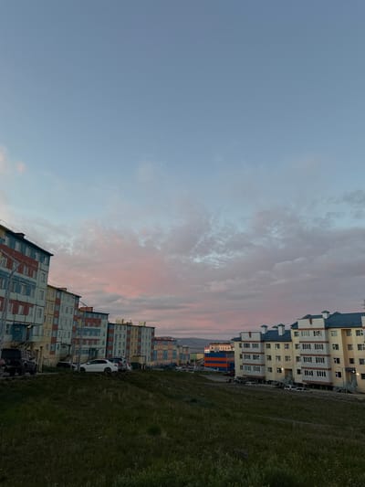 The image depicts a view of residential buildings in Anadyr, Russia, during what appears to be either dawn or dusk, given the soft pastel colors in the sky and clouds. The buildings are modern, multi-story apartment complexes with distinct color schemes, featuring sections of red, blue, yellow, and white facades. Some buildings have pitched roofs, while others are flatter. In the foreground, a grassy, sloped area is visible, with scattered patches of wildflowers. Several cars are parked along the street in front of the buildings. The overall impression is of a quiet, residential urban landscape under a colorful sky.