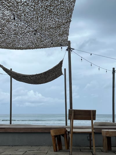 The image shows a patio with a view of the ocean in Las Escobas del Venado, Panama (latitude: 7.43, longitude: -80.188). The patio has a wooden deck with a wooden chair, wooden stool, and two hammocks strung with string lights. The chair is facing the ocean, and the stool is to the right of the chair. The ocean is in the background, and it is a light blue color. There are waves in the ocean. The sky is a light blue color with some white clouds. The hammocks are made of a beige material, and the string lights are strung between the two support poles. The string lights have five light bulbs.  The patio has a grey wall and stone pavers. The scene is empty of people.
