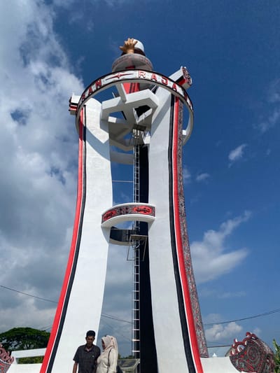 A low-angle, full-shot captures the "Tugu Selamat Datang" or Welcome Tower in Parlondut, Indonesia. The tower, a towering structure with a white base, is adorned with vertical bands of red, black, and white fabric featuring intricate cultural patterns. A large, circular band with red and white lettering encircles the upper portion of the tower, topped by a sculpture of a hand reaching upwards. Two individuals, a man and a woman wearing a hijab, stand at the base of the tower, seemingly observing the structure. The sky above is a vibrant blue with scattered white clouds, and a portion of a lush green tree is visible to the left of the frame. The tower exhibits traditional Indonesian motifs, suggesting a celebration of local heritage.