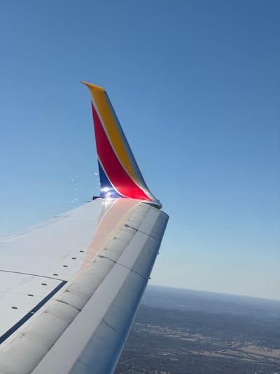 This image displays the port wing and winglet of a Southwest Airlines aircraft in flight, captured from a passenger's perspective. The winglet is painted with the airline's distinctive yellow, red, and blue livery, exhibiting a bright sun reflection where it joins the main wing structure. The aircraft wing, predominantly grey and white, shows visible panel lines and fasteners. The upper two-thirds of the frame consists of a clear, cloudless blue sky. Below the wing, a expansive landscape of rolling hills covered in dense, leafless deciduous trees extends to the horizon. Scattered light-colored buildings and visible road networks indicate a rural-to-suburban environment. The topographical features and visible development are consistent with the terrain surrounding Nashville, Tennessee, United States, suggesting the aircraft is in the vicinity of Nashville International Airport (BNA), likely during ascent or a banking maneuver. No individuals or direct interactions are visible.