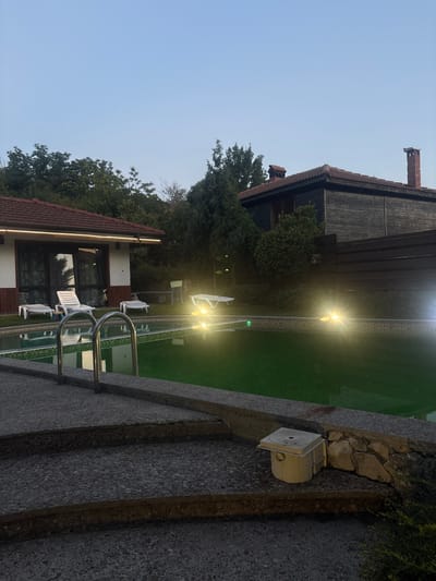 The image depicts a residential property with a swimming pool, likely located in Prohod, Bulgaria. The time appears to be dusk or early evening, as indicated by the soft natural light and the artificial illumination around the pool.
In the foreground, a concrete staircase leads down to the pool area. The pool itself is filled with green water and is illuminated by several bright lights placed around its perimeter. The pool has a tiled border. A metal ladder extends from the pool deck into the water. Adjacent to the pool is a small, light-colored utility box with a circular lid and several openings.
To the left of the pool is a single-story building with a white facade, dark-trimmed windows and doors, and a red-tiled roof. A string of lights runs along the edge of the building's roof. Several white lounge chairs are placed on a grassy area between the building and the pool.
In the background, a darker, wooden building with a red-tiled roof and a brick chimney is visible. This structure appears to be older and more rustic than the white building. Trees and dense vegetation surround the property, suggesting a somewhat rural or semi-rural setting within Prohod. The sky is a clear, pale blue, indicative of twilight. No people are present in the image.