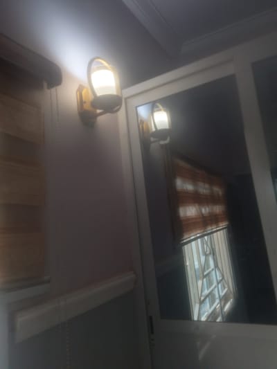 A blurred, indoor photograph shows two wall-mounted light fixtures. The one on the left is a vintage-style brass sconce with a white glass shade, casting a bright light upwards. To the right of the sconce, a vertical beaded chain hangs down the wall. A wooden slat blind is partially visible behind the chain. The second light fixture, on the right, is reflected in a mirror. This fixture also appears to be a brass sconce with a white glass shade, and it is mounted next to a striped brown and white blind. The mirror also reflects a window with a criss-cross metal security grate. The overall impression is that of an interior space, possibly a hallway or room, in Gboko, Nigeria.