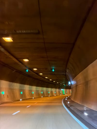 The image shows the interior of a road tunnel, presumably in Farchant, Germany. The tunnel has a curved structure with a light brown, textured ceiling and walls. The road surface is paved and marked with white lane lines. Along the walls and ceiling, there are numerous lights, some appear to be rectangular and yellow, while others are smaller and green, some of which are in the shape of arrows. There is also a blue square sign with a white symbol visible on the right side of the tunnel wall. The lighting creates a warm, ambient glow, with reflections visible on the road. The perspective suggests the photo was taken from a moving vehicle, looking forward into the tunnel. There are no people or other vehicles clearly visible in the image. The environment is enclosed and artificial, typical of an infrastructure tunnel.