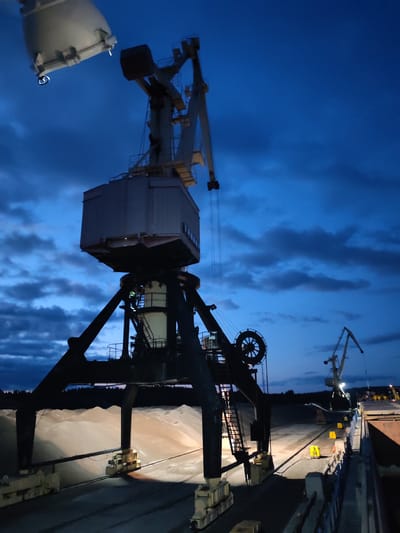 The image depicts a nighttime scene at a port or industrial facility in Shalsky, Russia. Two large cranes dominate the foreground, one much closer and more detailed than the other in the background. The closer crane, a bulk handling machine, is a dark silhouette against a deep blue, cloudy sky. It features a large cabin structure at its top, supported by a complex of beams and supports. A large clam-shell bucket or similar grab mechanism is suspended from the boom of this crane.
To the left of the main crane, a second, lighter-colored crane structure is partially visible, extending upwards into the frame. In the distance, a smaller, lighter-colored crane with a long boom is also visible, its outline defined by lights.
The ground area in front of the main crane is piled high with a light-colored granular material, possibly sand or aggregate, illuminated by spotlights. Railway tracks or a similar transport system are visible on the ground, with yellow markers placed along the edges of the material piles. A ship or large barge is docked to the right, its hull visible as a dark structure. The overall impression is one of industrial activity operating at night, with artificial lighting playing a key role in defining the scene. There are no visible people in the image.