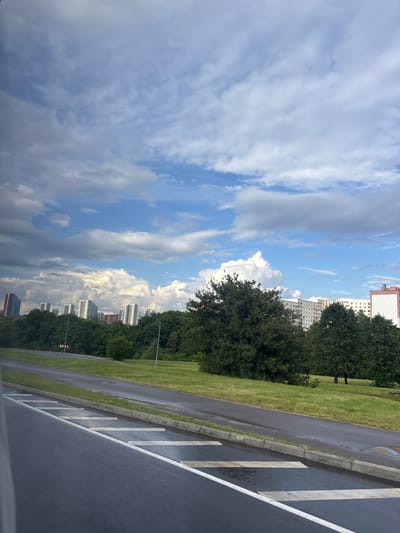 The image depicts a cityscape with residential buildings visible in the background, partially obscured by lush green trees. The foreground features a wet asphalt road with distinct white lane markings, suggesting recent rain. The sky is a mix of blue and dramatic grey clouds, indicating potentially changing weather conditions. The scene appears to be an urban or suburban environment within the Сеніцкі сельскі Савет, Belarus, characterized by a blend of natural greenery and modern architecture.