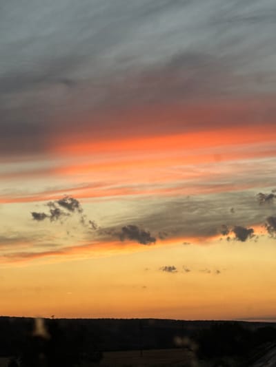 This is a vertical, eye-level photograph of a sunset over a rural landscape in Veselina, Bulgaria. The sky is filled with dramatic clouds displaying a gradient of colors from dark gray at the top to fiery orange and soft yellow near the horizon. Wispy streaks of pink and orange are visible across the middle of the sky, illuminated by the setting sun. Below the colorful sky, a dark silhouette of rolling hills and trees forms the horizon line. The foreground is also dark, with indistinct shapes suggesting trees and possibly some vegetation. The overall impression is one of natural beauty and a peaceful evening scene.
