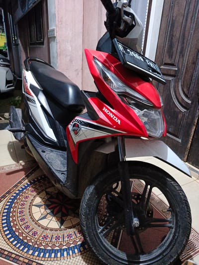 A red and white Honda scooter is parked on a patterned tiled floor in front of a wooden door and a pink-toned wall. The scooter has a black seat and black handlebars. A license plate that reads "B 3900 FWI" is attached to the front. The scooter has a white front fender and a red body with white and black graphics, including "HONDA" written in white on the front fairing. The scooter is the primary subject of the image. The environment appears to be an outdoor, covered area, possibly a porch or carport, with a building facade visible in the background. No people are present in the image. The location is specified as Payacut, Indonesia.